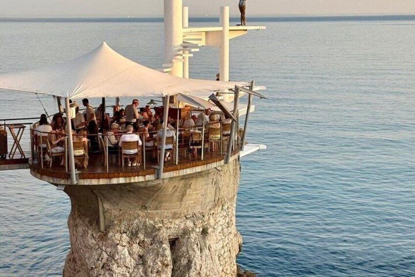 A dining area on a cliff overlooking the sea, with a white structure in the background. 2
