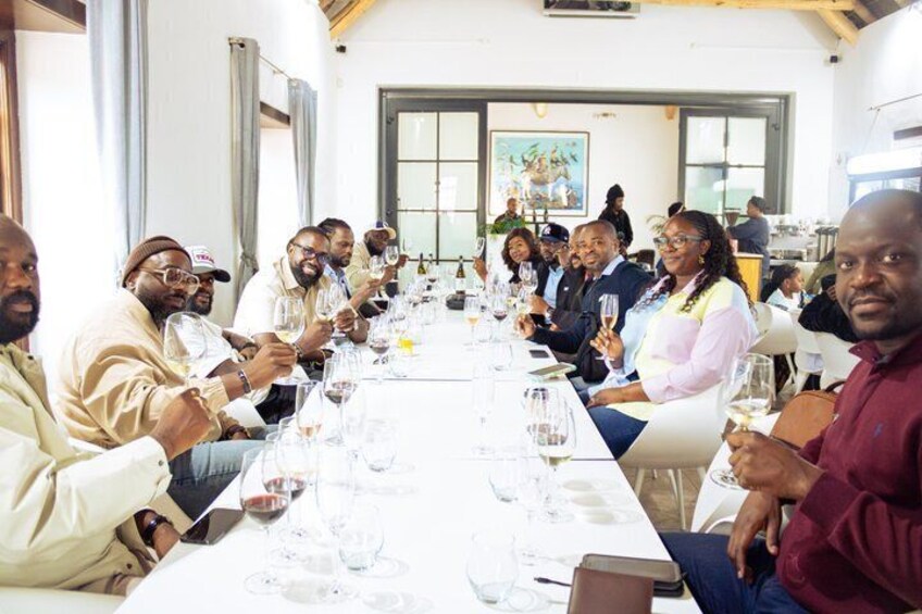 A group of people seated at a long table with wine glasses, in a room with large windows and a painting on the wall. 3