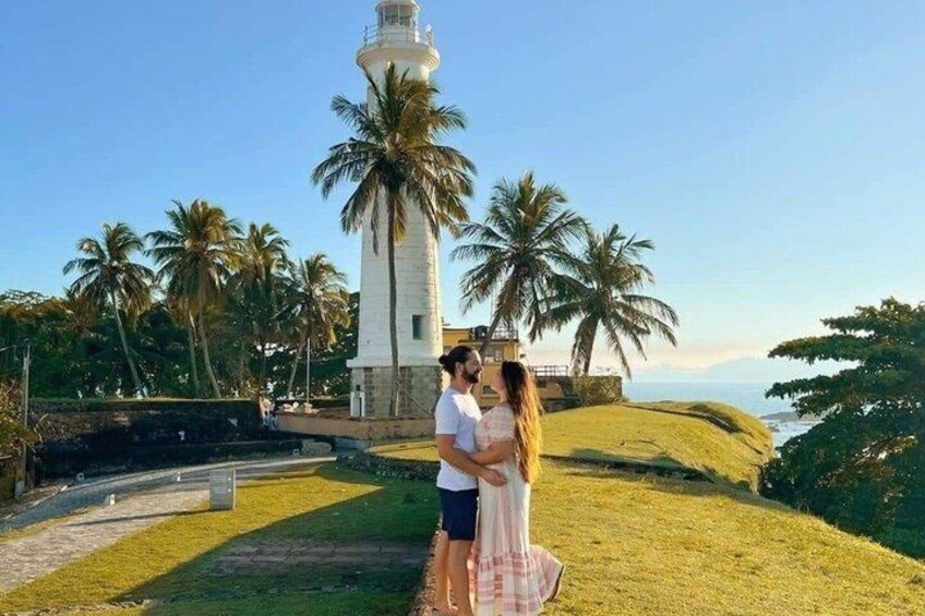 A couple embracing near a lighthouse with palm trees and a grassy hill in the background. 4
