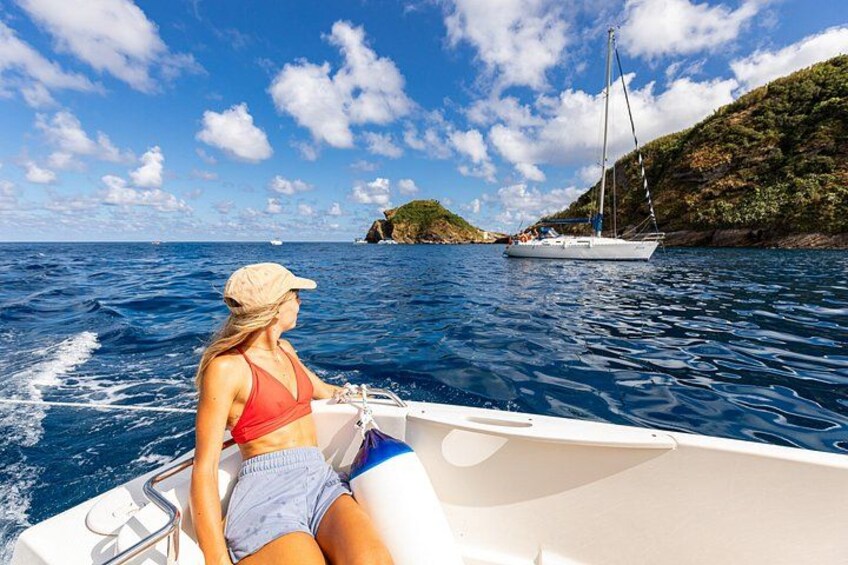 A woman on a boat, a sailboat in the background, and a rocky coastline. 3