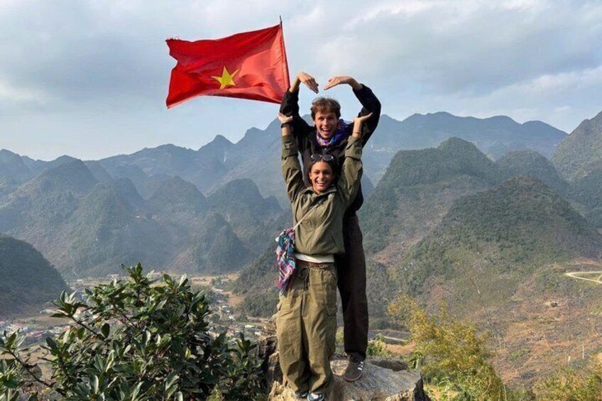 Two people standing on a mountain with a flag, surrounded by a mountainous landscape. 1