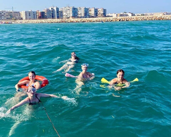 People in the water with floatation devices, a rocky shoreline, and buildings in the background. 3