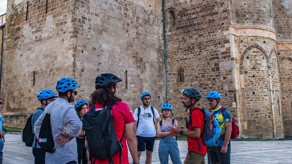 A group of people wearing helmets and backpacks standing in front of a historic stone building with arched windows and a circular tower. 4