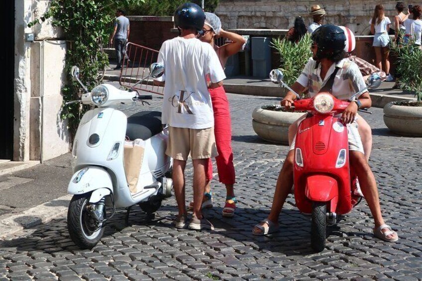 A cobblestone street with two people on scooters, one white and one red, and a person standing nearby. 3