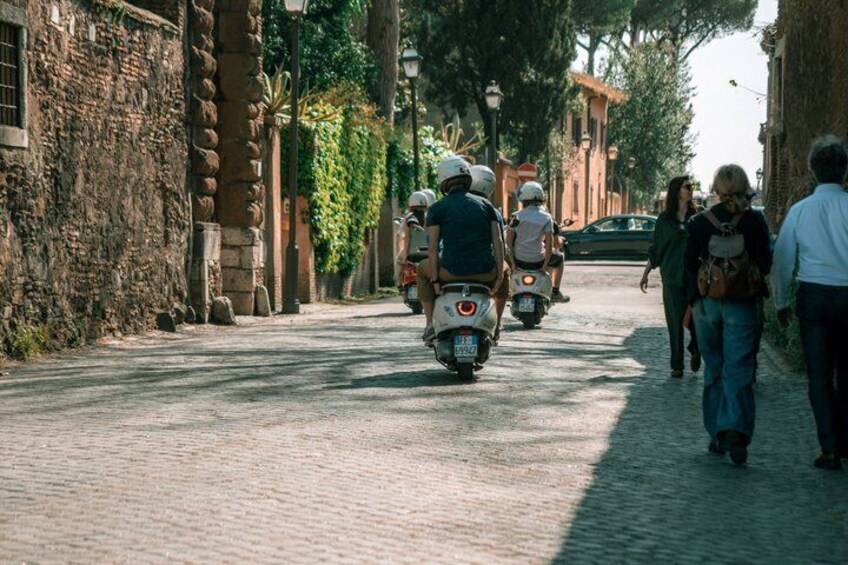 A cobblestone street with people walking and motorcyclists riding. 1