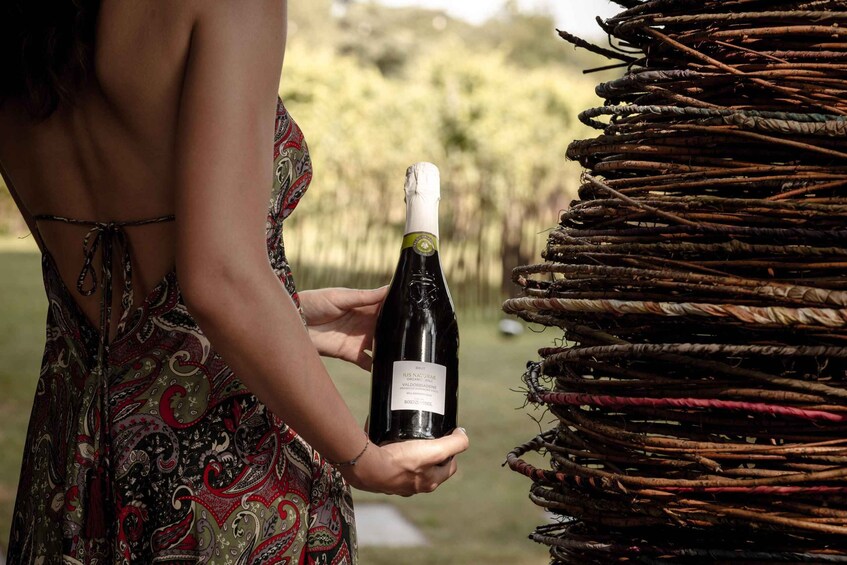 A person holding a bottle of wine with a label, standing next to a stack of woven baskets. 2