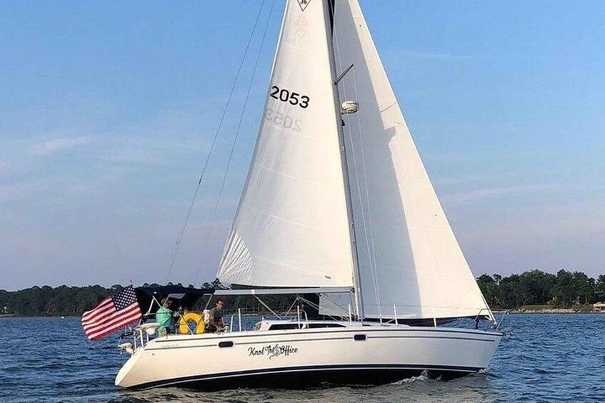 A sailboat with the number 2053 on its sail, named 'Knot the Office,' on a body of water with an American flag and a person visible on the deck. 1