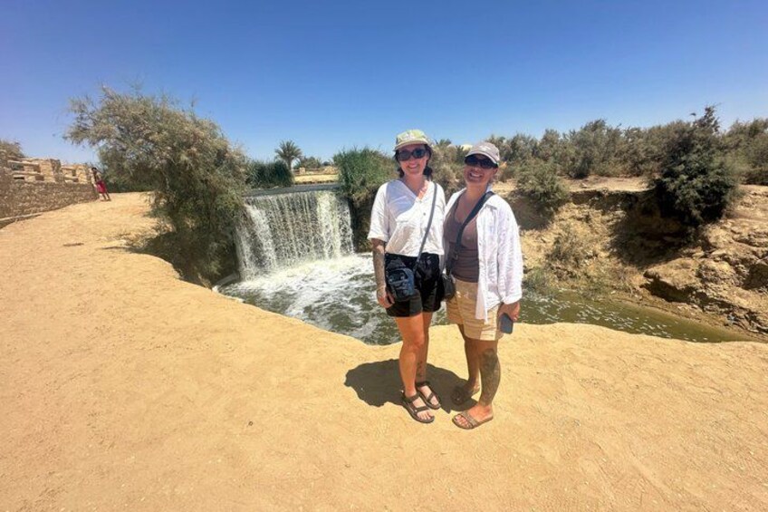 Two people standing by a small waterfall in a desert setting with sparse vegetation. 5