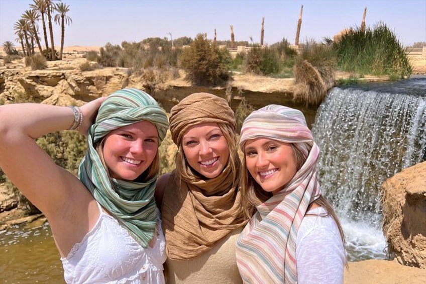 Three people standing by a waterfall in a desert landscape with palm trees and rocky terrain. 5