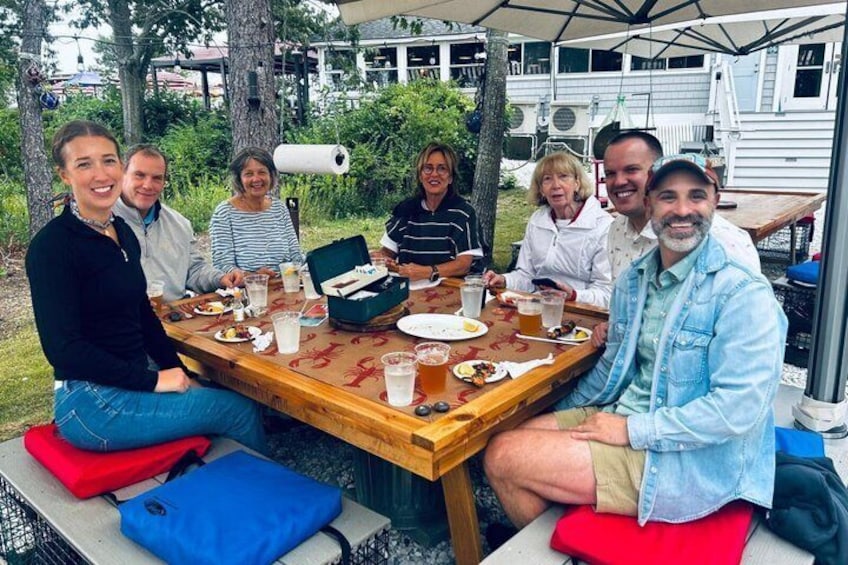 A group of people sitting around a wooden table outdoors with food and drinks. 2