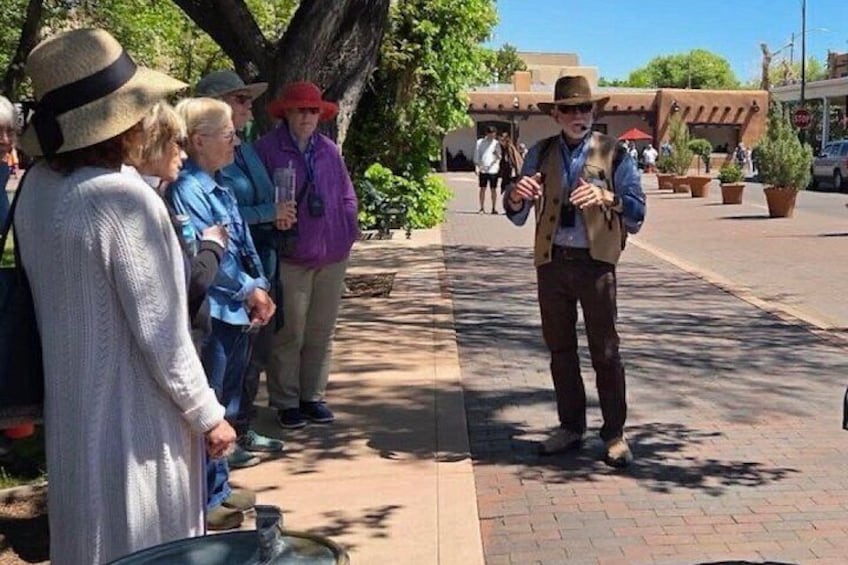 A person in a hat and vest speaking to a group of people on a sidewalk. 1
