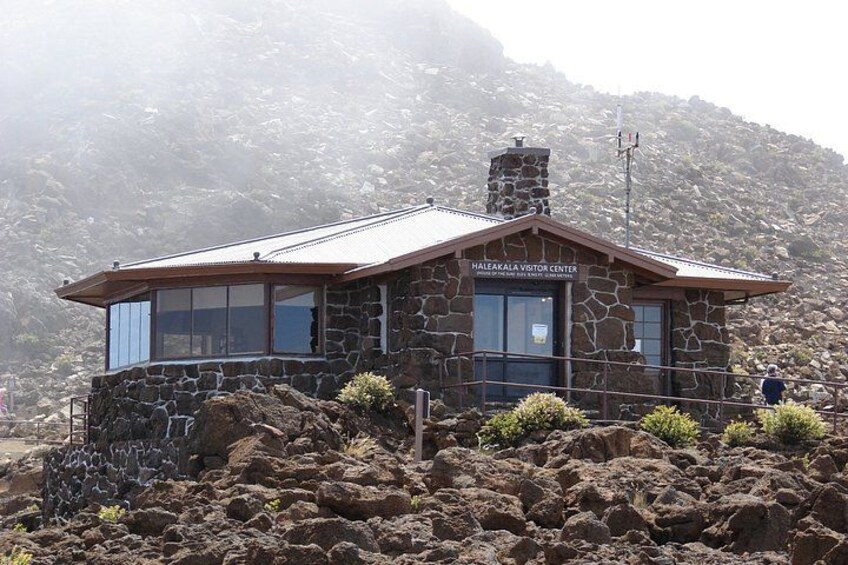 A stone building with a metal roof, labeled 'Haleakala Visitor Center,' surrounded by rocky terrain and a mountainous backdrop. 3