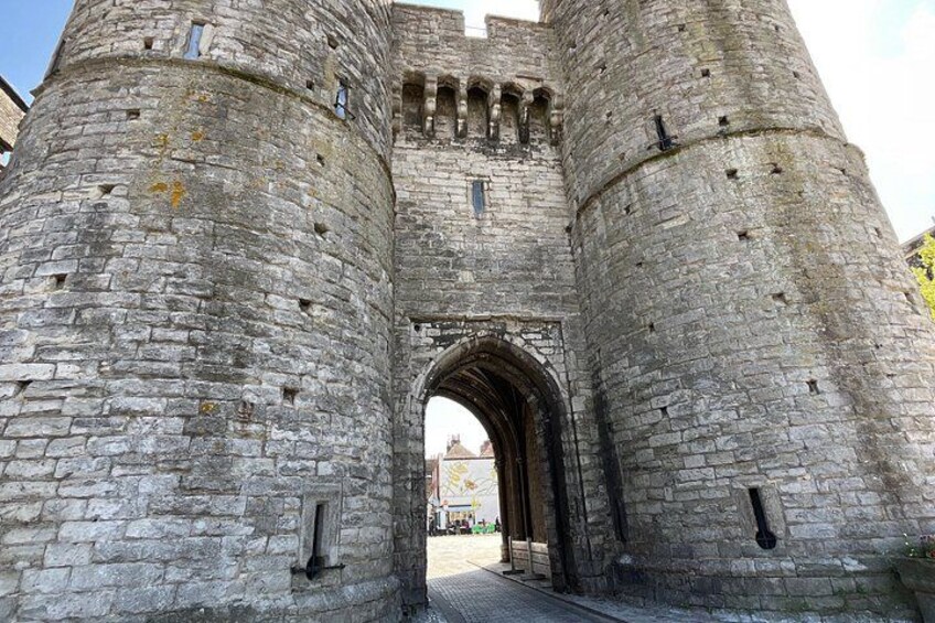 A medieval stone castle gate with a large archway, flanked by two tall towers with battlements. 1