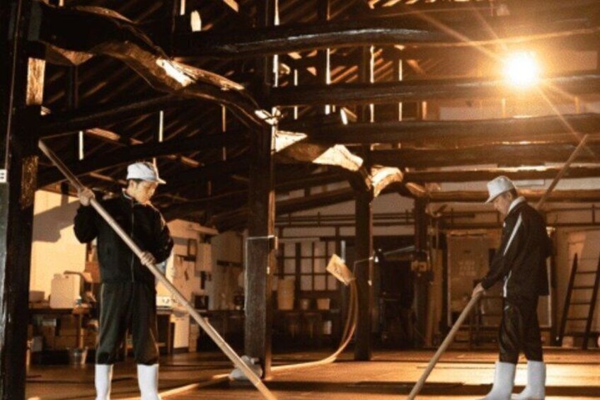 Two workers in a dimly lit industrial setting, using long-handled tools to manage materials. 1