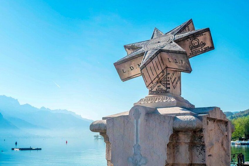 A cross-shaped monument with inscriptions, a body of water, and mountains in the background. 5