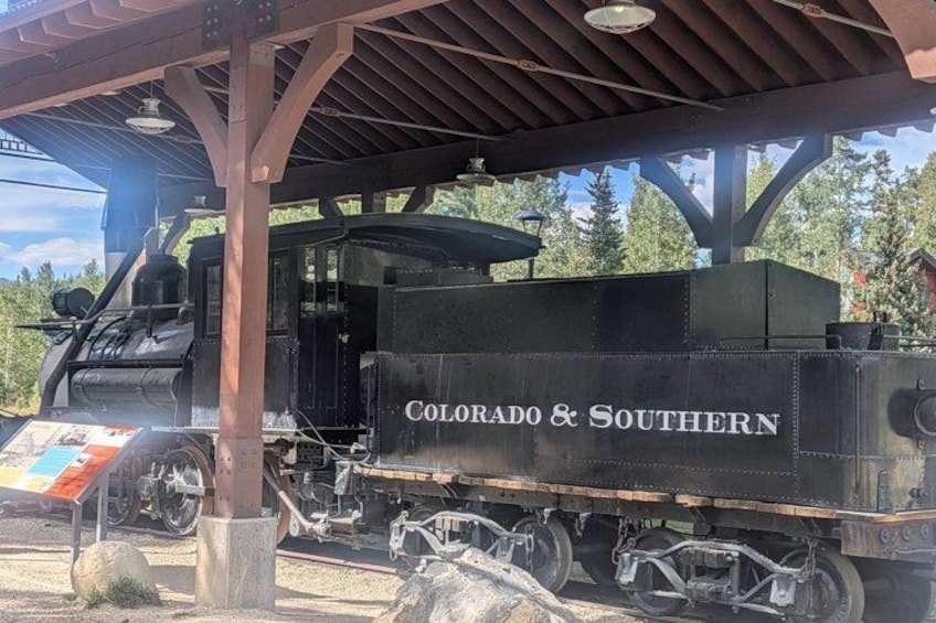 A steam locomotive named 'Colorado & Southern' parked under a covered area with a display board nearby. 5