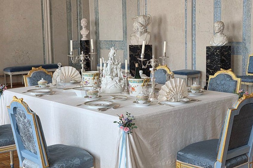A formal dining table set with white tablecloth, blue upholstered chairs, and classical bust sculptures. 4