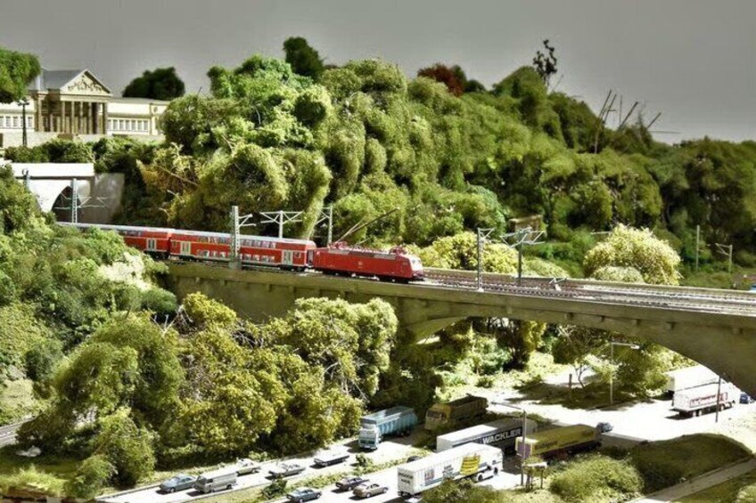 A model train on a bridge, surrounded by greenery and a building. 1