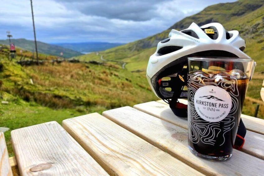 A bicycle helmet and a glass of KirKStone Pass beer on a wooden table with a scenic view of hills in the background. 4