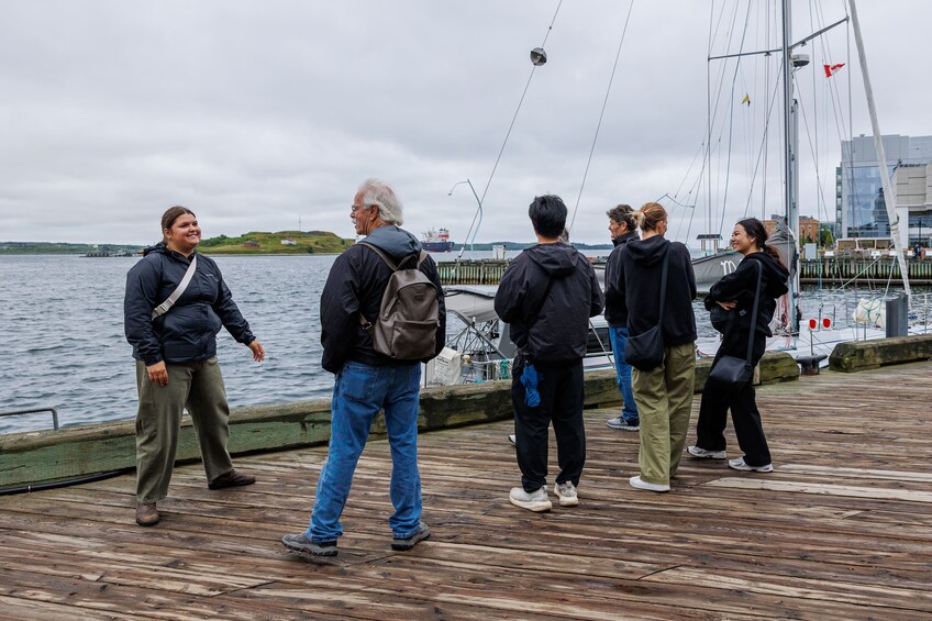 Eine Gruppe von Menschen steht an einem hölzernen Steg neben einem Gewässer, im Hintergrund ein Segelboot und eine Stadtansicht. 2