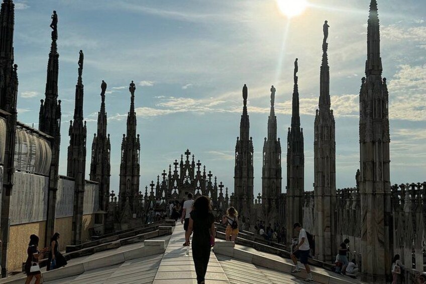 A Gothic-style cathedral with tall spires and a central dome, a paved walkway, and people walking and sitting. 1