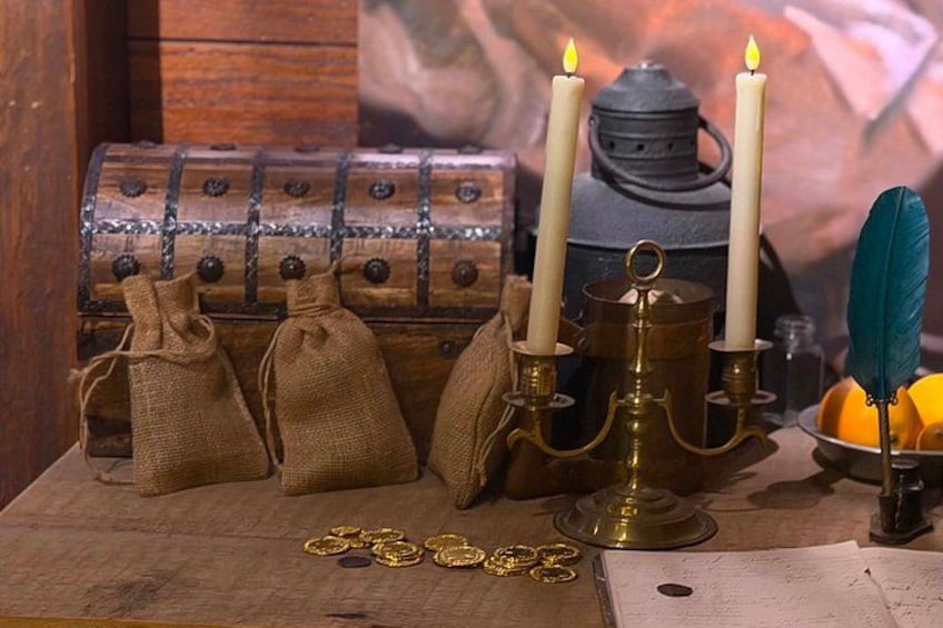 A wooden chest, three candles, a feather quill, and some gold coins on a table. 4