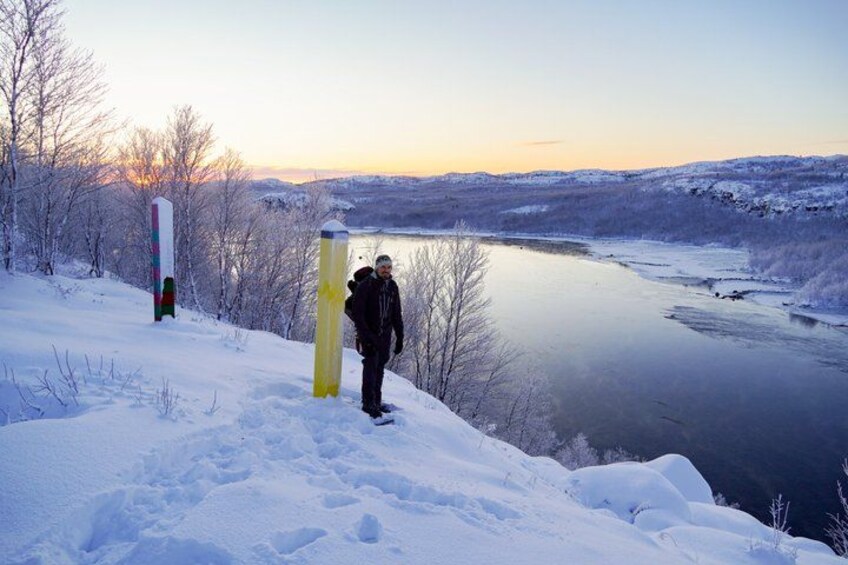 A snow-covered landscape with a river, a person standing near a yellow object, and a signpost in the background. 1