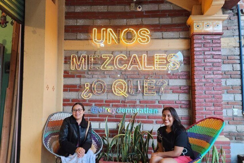 Two people sitting on wicker chairs in front of a brick wall with a neon sign. 1