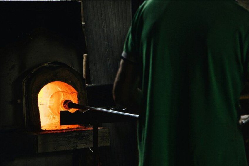 A person in a green shirt working with a long metal rod in front of a glowing furnace. 3