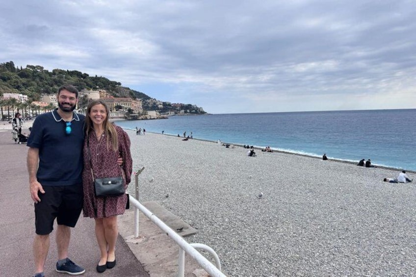 A couple standing on a pebbled beach with a railing, a clear sky, and buildings in the background. 2