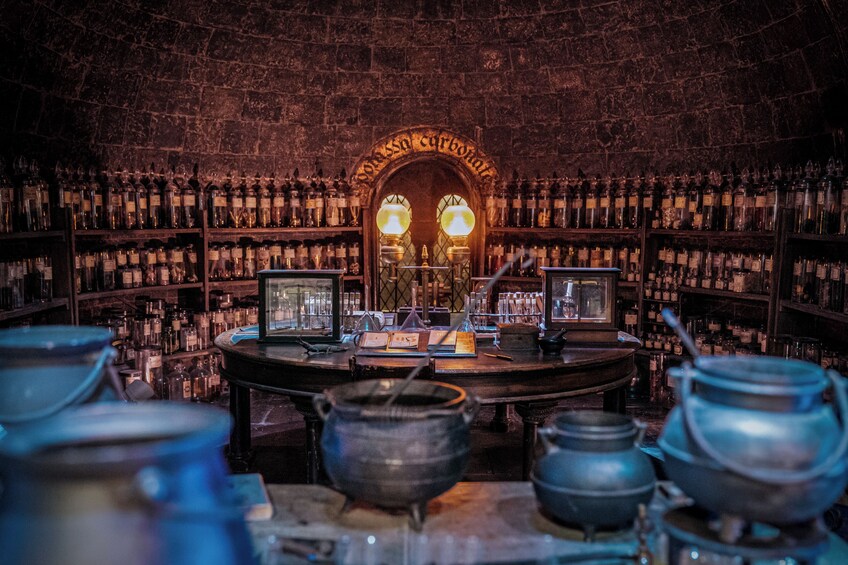 A dimly lit room with shelves of glass bottles and jars, a central table with various objects, and a stone wall with an arched doorway. 2