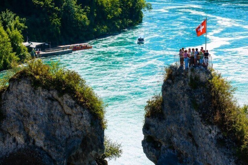 A rocky cliff with a Swiss flag, a boat on the river, and a group of people on the cliff. 5