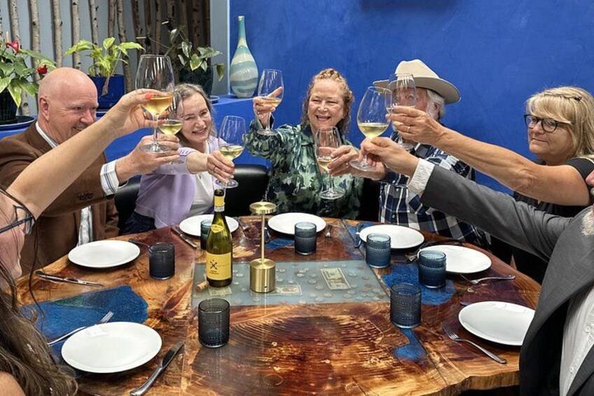 A group of people toasting with wine glasses around a wooden table with plates, cutlery, and a bottle. 5