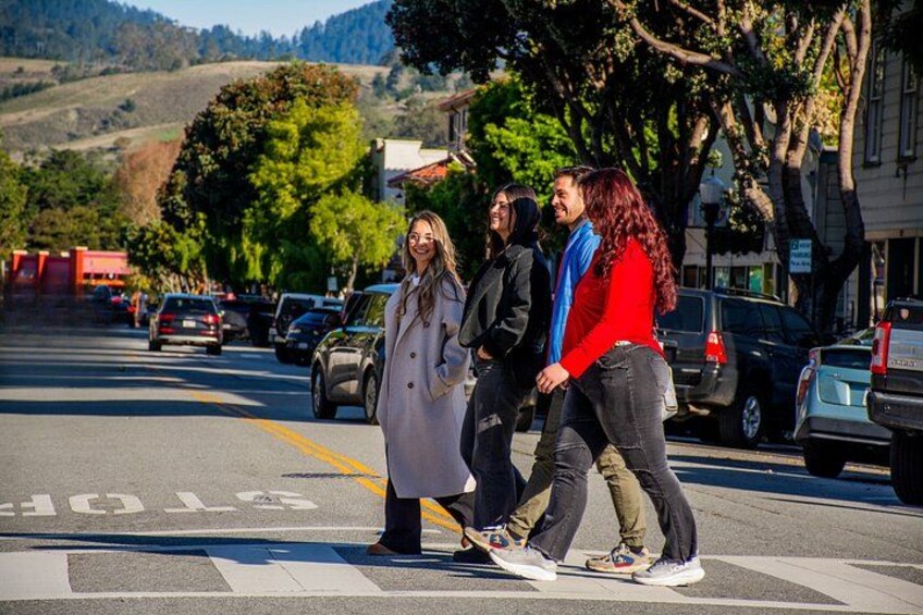 A street scene with pedestrians crossing, parked cars, and a hilly landscape in the background. 3