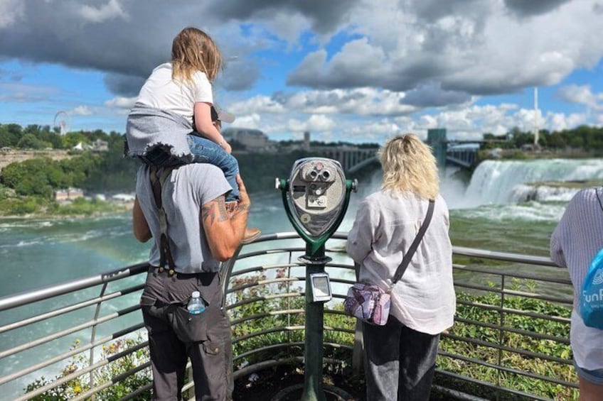 A person carrying a child on their shoulders, another person using binoculars, and a waterfall in the background. 1