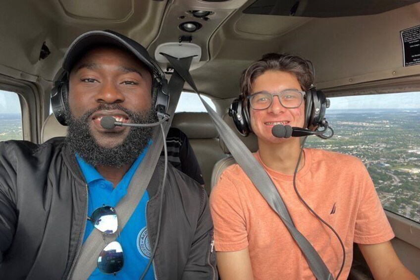Two people in a helicopter cockpit with a view of a landscape below. 1