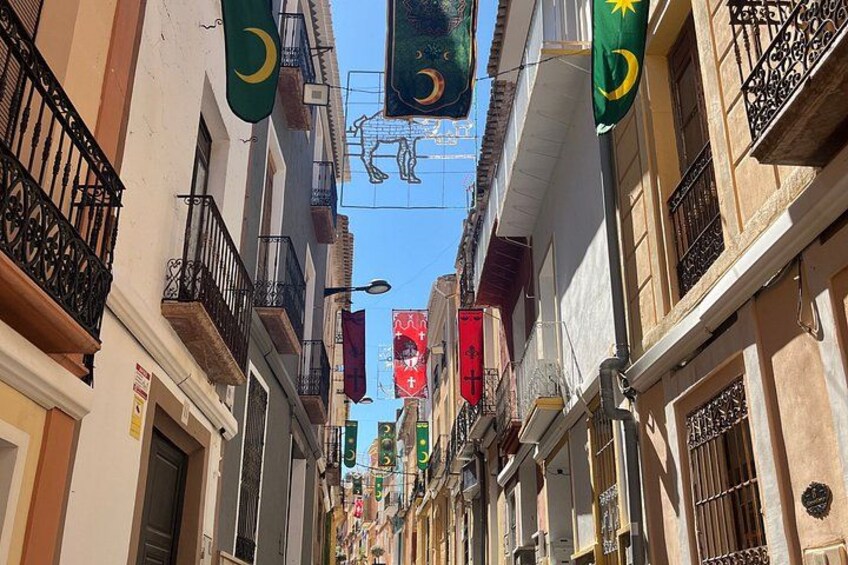 A narrow street with buildings, flags, and balconies. 2