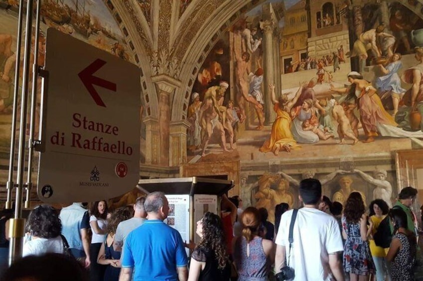 A crowded museum with a frescoed ceiling and a sign pointing to 'Stanze di Raffaello.' 4