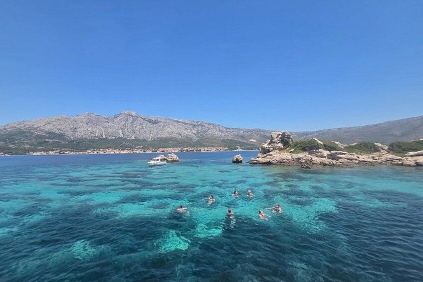 A clear blue sea with people swimming and boats in the distance, rocky coastline, and mountains in the background. 2