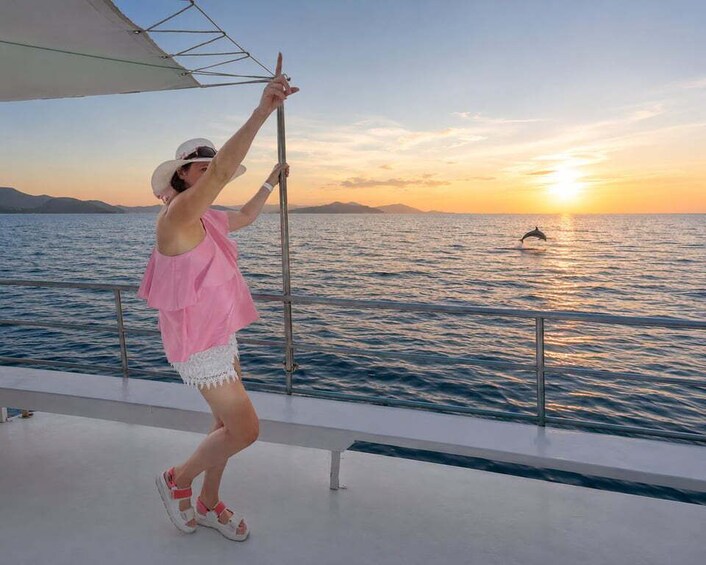 A person on a boat deck, holding onto a railing, with a sunset and dolphin in the background. 2