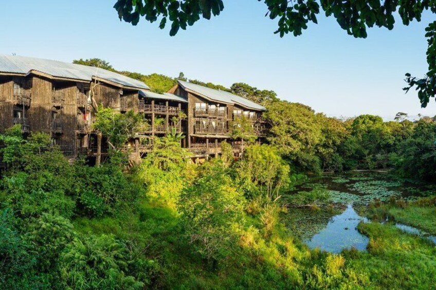 A wooden house with a balcony, surrounded by lush greenery and a pond. 2