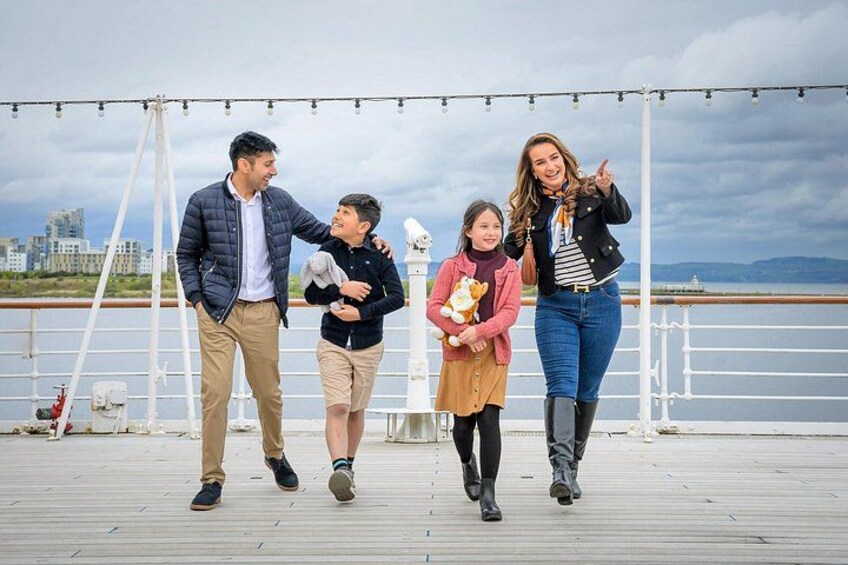 A group of people walking on a wooden deck by a body of water with a cityscape in the background. 2