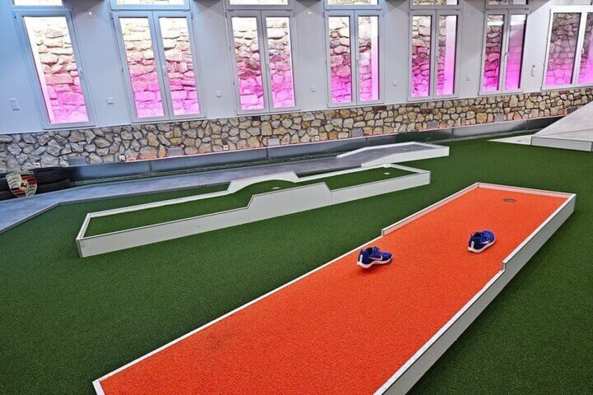 A miniature golf course with a red and green playing surface, stone wall, and windows with pink lighting. 5
