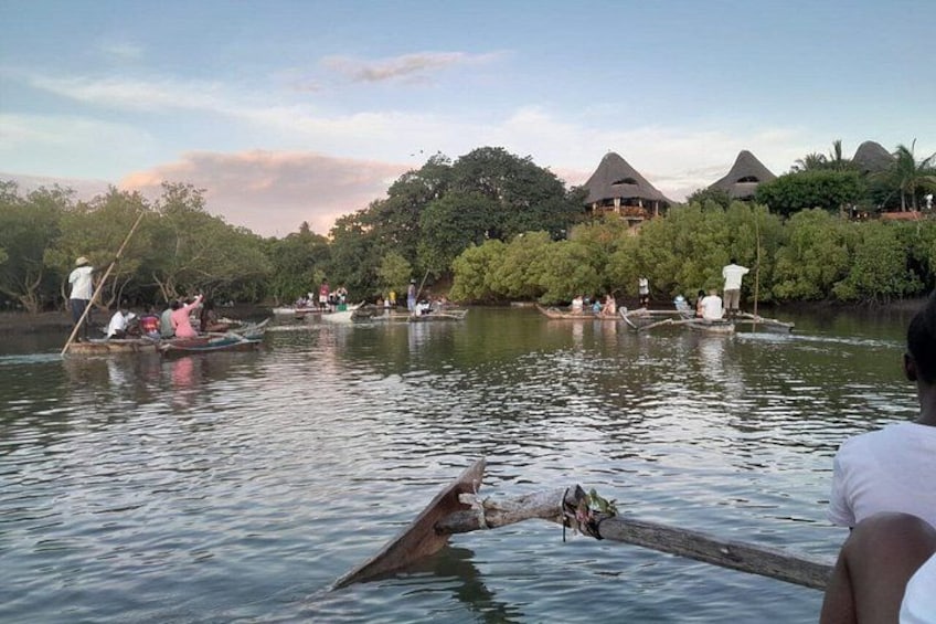 A river with people in boats, a person paddling, and thatched-roof buildings in the background. 3