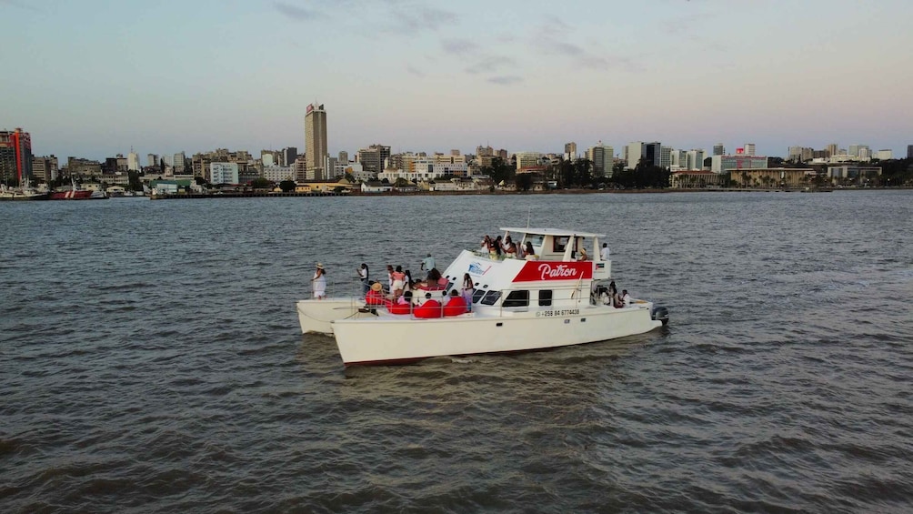 A white boat with red accents named 'Patron' on a body of water with a city skyline in the background. 1