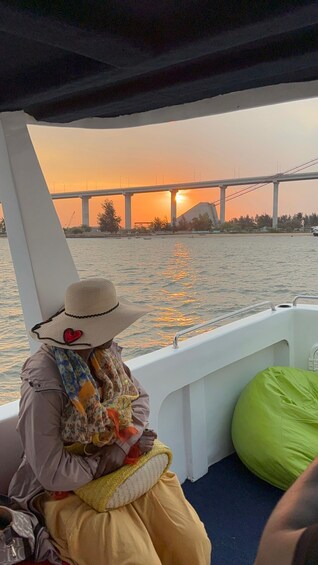 A person on a boat, wearing a hat and a floral scarf, looking at a sunset over a bridge. 2