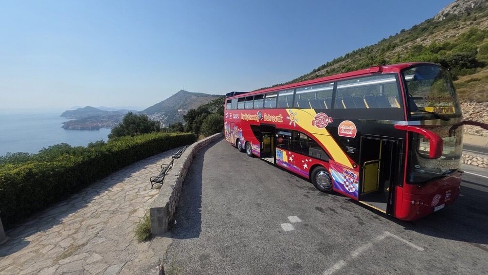 En rød toetagebus parkeret på en snoet vej med et smukt udsigt over havet og bakkerne i baggrunden. 3