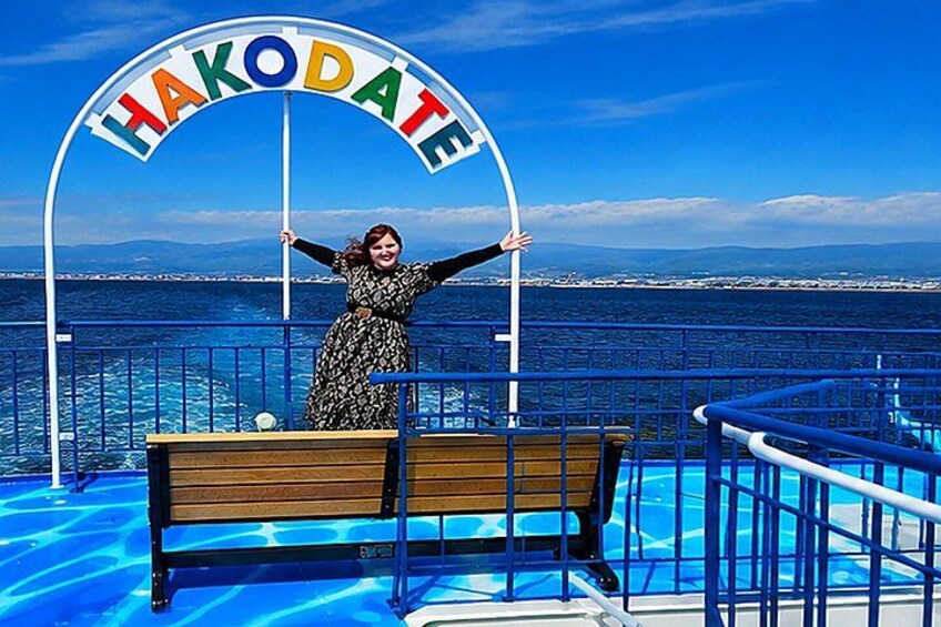 A person standing on a deck with a sign that reads 'HAKODATE' and a view of the sea and mountains in the background. 3