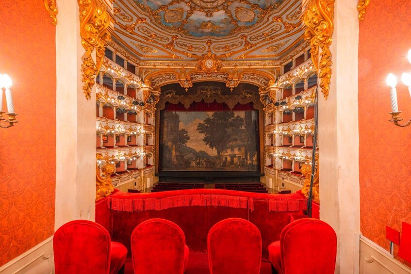 An ornate theater interior with red velvet seats, gold decorations, and a large painting on the back wall. 3