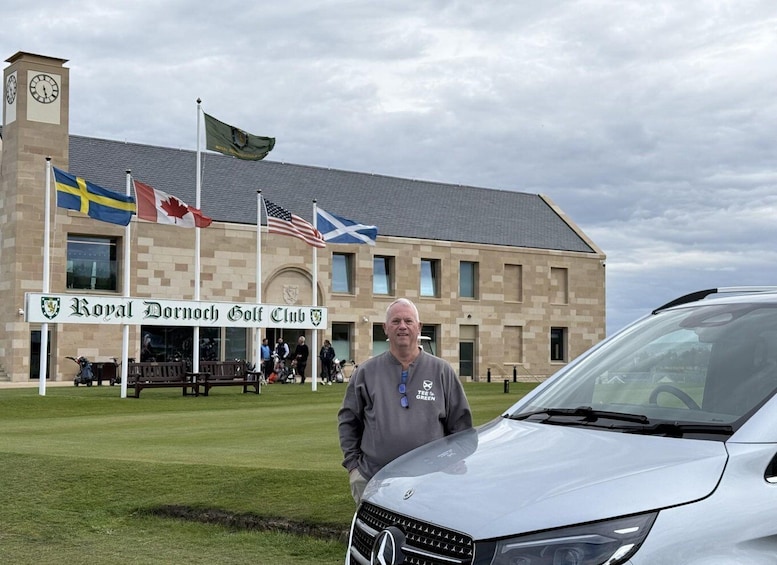 A man standing in front of a building with flags, a golf club sign, and a car. 1